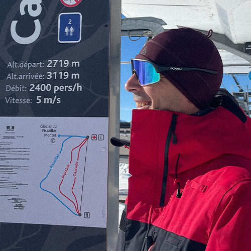 Person in red jacket and sunglasses standing next to a ski lift information sign